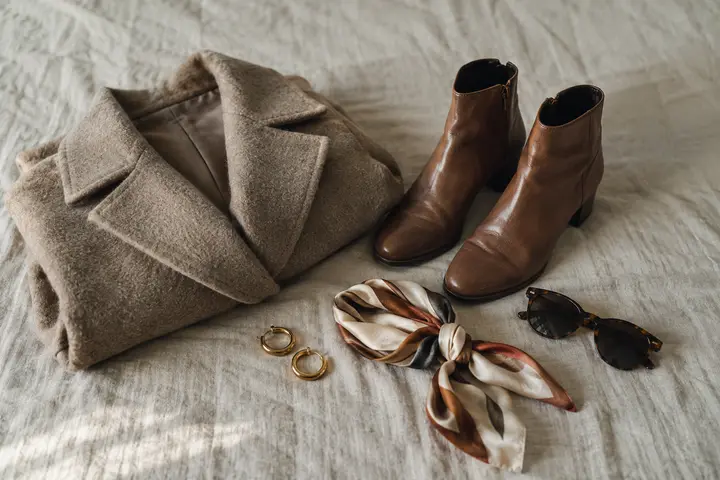 A wool coat, leather boots, gold earrings, a silk scarf and sunglasses laid out on linen.