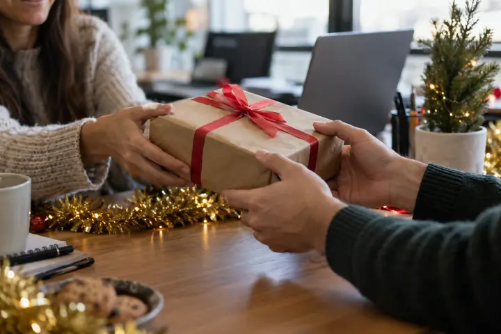 Coworkers passing a wrapped gift across a festive office desk.