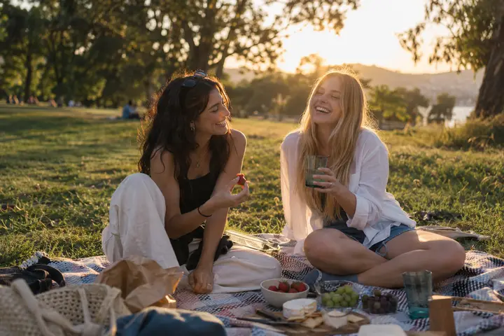 Two friends laughing on a picnic blanket in a park.
