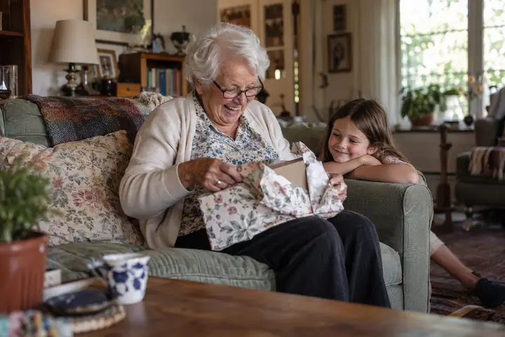 A grandmother laughing as she unwraps a gift while her granddaughter watches.